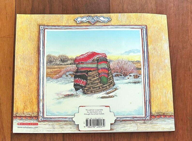 May include: The back cover of a children's book shows a winter scene with a woven basket filled with colourful blankets and scarves. The illustration is framed with a wooden border and a decorative element at the top.