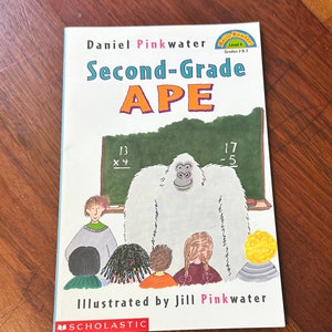 Puede incluir: Una portada de libro infantil con un yeti blanco de pie frente a una pizarra con problemas de matemáticas. El título del libro es "Second-Grade APE" de Daniel Pinkwater, ilustrado por Jill Pinkwater. El libro es publicado por Scholastic.