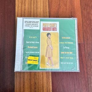 May include: A CD case with a green background and a picture of Patsy Cline. The title of the CD is "Patsy Cline's Greatest Hits". The CD case has a yellow sticker that says "Best Buy" and "$9.92".