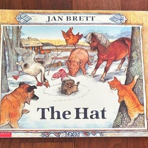 May include: A children's book cover for "The Hat" by Jan Brett. The illustration shows a snowy farmyard with a goose, dog, pig, horse, cat, and chicken. The book is framed with a wooden border.