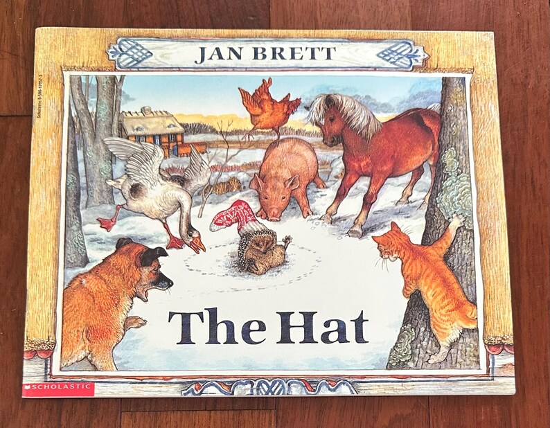 May include: A children's book cover for "The Hat" by Jan Brett. The illustration shows a snowy farmyard with a goose, dog, pig, horse, cat, and chicken. The book is framed with a wooden border.