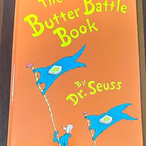 Könnte beinhalten: Ein orangefarbenes Hardcover-Buch mit dem Titel "The Butter Battle Book" von Dr. Seuss. Das Cover zeigt gelben Text und Illustrationen von blauen Flaggen mit Butterbrot. Ein Kinderbuch.
