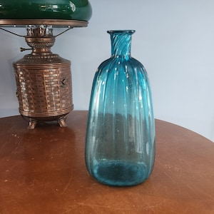 May include: A blue glass bottle with a ribbed design sits on a wooden table. The bottle is empty and has a rounded bottom.