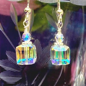 May include: A pair of gold-toned earrings with iridescent, rectangular crystal pendants. Each earring features a small, round crystal above the pendant and a gold-colored hook. The crystals reflect a spectrum of colors, including blue, green, and yellow.