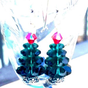 May include: A pair of Christmas tree earrings. Each earring features a dark green, faceted bead tree design topped with a pink and red crystal. The earrings have silver-colored decorative bases and ear wires, perfect for holiday wear.