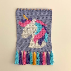 Puede incluir: Un tapiz de pared tejido a crochet con un unicornio con una melena y un cuerno arcoíris. El unicornio está sobre un fondo morado con un borde de flecos.