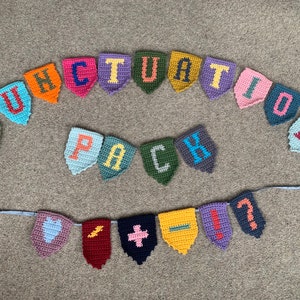 Può includere: Uno striscione all'uncinetto con le parole "Punctuation Pack" e vari segni di punteggiatura. Lo striscione è realizzato con filati di diversi colori e presenta un cuore, un fulmine, un segno più, un segno meno, un punto esclamativo e un punto interrogativo.