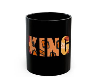 Puede incluir: Taza de cerámica negra con la palabra "KING" en letras estampadas en naranja, marrón y amarillo. La taza es cilíndrica y tiene un acabado brillante. El diseño está centrado en la taza.