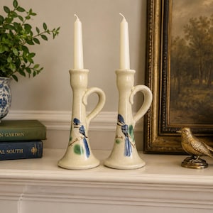 May include: Two cream-colored ceramic candle holders with handles, each featuring a hand-painted blue bird perched on a branch. The holders have a speckled finish and are each topped with a tall, slender, off-white taper candle.