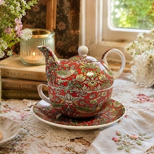 May include: A red and green floral patterned tea-for-one set. The set includes a teapot, teacup, and saucer. The teapot is stacked on the teacup, which sits on the saucer. The set is on a floral tablecloth, with a plate of cookies and a vase of flowers nearby.