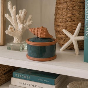 May include: A round, dark teal ceramic lidded jar with a terracotta-colored rim and base. The lid features a sculpted, light brown seashell handle. The jar is positioned on a shelf alongside a white coral sculpture and a white starfish.