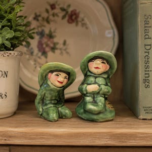 May include: Two ceramic figurines depicting figures wearing green hats and matching green outfits. The figures have painted facial features, including red lips and dark hair. One figure is in a seated position, while the other is kneeling.