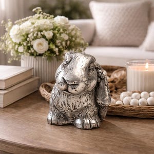 May include: A silver-colored rabbit figurine with long, floppy ears sits on a wooden coffee table. A lit candle in a glass jar, a bouquet of white flowers, and two books are also on the table. The scene is set in a living room.