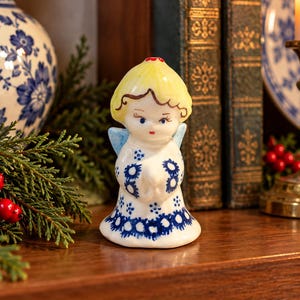 May include: A ceramic angel figurine with yellow hair, blue wings, and a white dress featuring a blue floral pattern. The angel has its hands clasped together in front of its chest, with painted facial features including eyes and red lips.