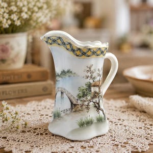 May include: A white porcelain pitcher featuring a hand-painted landscape scene with a bridge, trees, and a small building. The rim is decorated with a yellow and blue lattice pattern. The pitcher has a curved handle and a scalloped base.