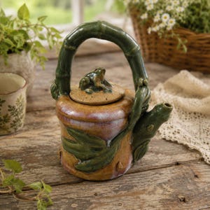 May include: A ceramic teapot featuring a tan body with a glossy finish, adorned with raised green leaf motifs. The handle is shaped like a bamboo stalk, and the lid is topped with a small, sculpted frog figure. The spout is also green.