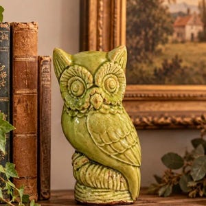 May include: A ceramic owl figurine featuring a glossy, light green glaze with a visible crackle finish. The owl is depicted perched on a textured, cylindrical base, showcasing detailed feather patterns, large circular eyes, and pointed ear tufts.