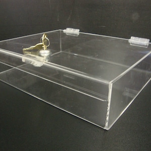May include: A clear acrylic lockable display box with a key and lock in the lid. The box is rectangular and has a hinged lid.