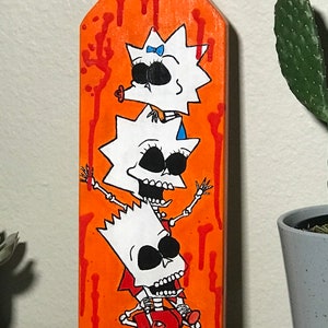May include: A wooden paddle painted orange with red splatters and three cartoon skeletons of the Simpsons characters, Bart, Lisa, and Maggie. The skeletons are stacked on top of each other with Maggie on the bottom, Lisa in the middle, and Bart on top.
