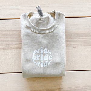 May include: A light beige sweatshirt with the words "bride bride bride" in a white circle design. The sweatshirt has a tag that says "miellis studio".