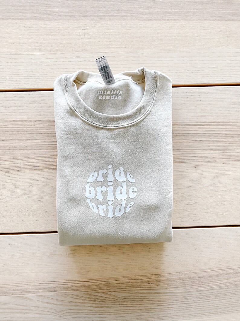 May include: A light beige sweatshirt with the words "bride bride bride" in a white circle design. The sweatshirt has a tag that says "miellis studio".