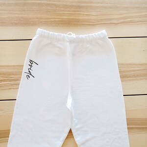 May include: White sweatpants with the word "bride" written in black on the left leg.