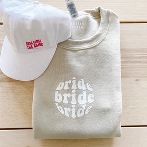 May include: A beige sweatshirt with the word "bride" written three times in a circle in a retro font. A white baseball cap with the text "Here comes the bride" in red is also pictured.