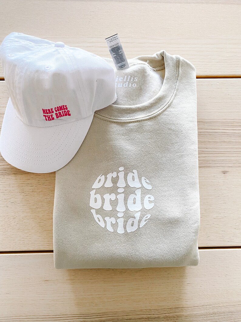 May include: A beige sweatshirt with the word "bride" written three times in a circle in a retro font. A white baseball cap with the text "Here comes the bride" in red is also pictured.