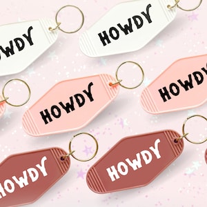 May include: A set of six keychains with the word "HOWDY" in a retro font. The keychains are in three colors: white, pink, and brown. Each keychain has a gold metal ring.