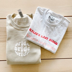 May include: Two beige sweatshirts with white text. One sweatshirt says "bride bride bride" in a retro font. The other sweatshirt says "Kylie's Last Disco" in pink and red text.