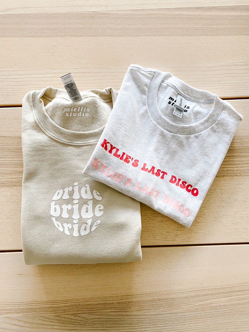 May include: Two beige sweatshirts with white text. One sweatshirt says "bride bride bride" in a retro font. The other sweatshirt says "Kylie's Last Disco" in pink and red text.