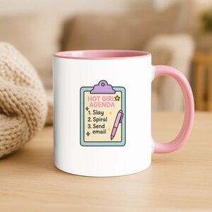 May include: A white ceramic mug with a pink interior and handle. The mug features a colorful illustration of a clipboard with the text "HOT GIRL AGENDA" and a list of tasks. A pink pen is also shown.