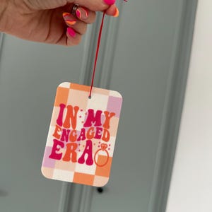 May include: A pink and orange chequered car air freshener with the text "In My Engaged Era" in pink and white letters. A diamond ring is drawn next to the text.