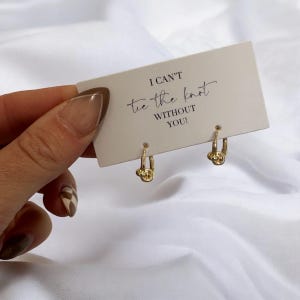 Puede incluir: Un par de pendientes de nudo de oro con una tarjeta que dice "No puedo atar el nudo sin ti!"