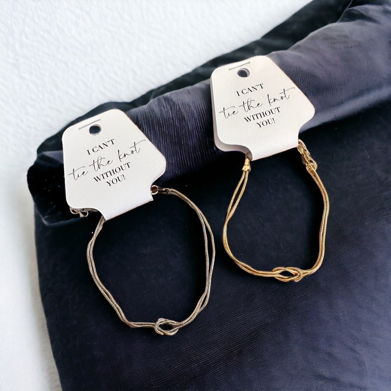 May include: Two gold-colored bracelets with a knot design are displayed on a dark background. Each bracelet is attached to a white tag that reads "I CAN'T tie the knot WITHOUT YOU!" in black script.
