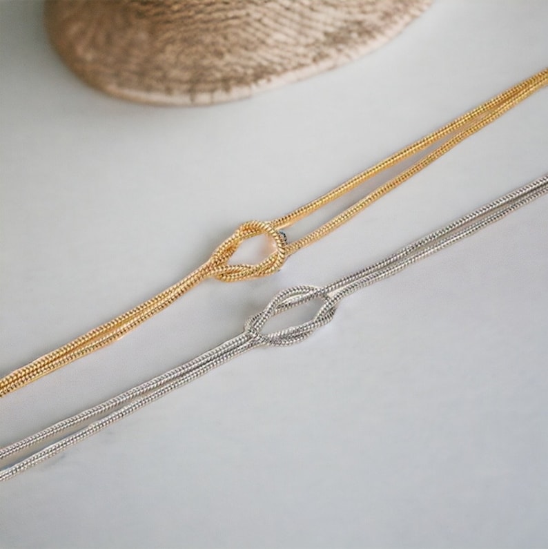 May include: Two delicate necklaces, one gold and one silver, each featuring a knot design. The necklaces are displayed on a light gray surface, with a woven texture in the background. The chains are thin and elegant.