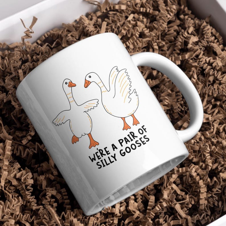 May include: White ceramic mug featuring two cartoon geese with orange feet and beaks. The mug has the text "WE'RE A PAIR OF SILLY GOOSES" in black. The mug is inside a white box with brown paper filler.