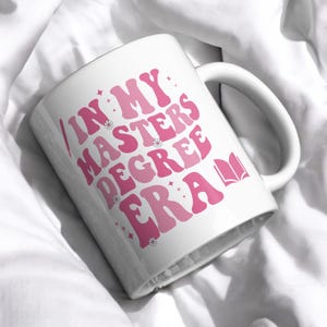 May include: White ceramic mug with a pink design that says "In My Masters Degree Era" with a pink book icon.