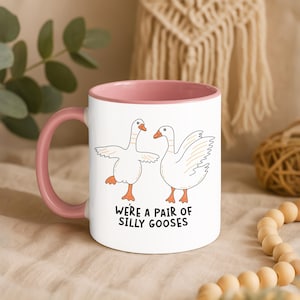 May include: White ceramic mug with a pink interior and handle. The mug features a cartoon illustration of two geese with orange feet and beaks. The text "WE'RE A PAIR OF SILLY GOOSES" is printed below the geese.