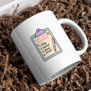 May include: White ceramic mug with a handle, featuring a colorful design. The design includes the text "HOT GIRL AGENDA" above a list: "1. Slay, 2. Spiral, 3. Send email." A pink pen illustration is next to the list.