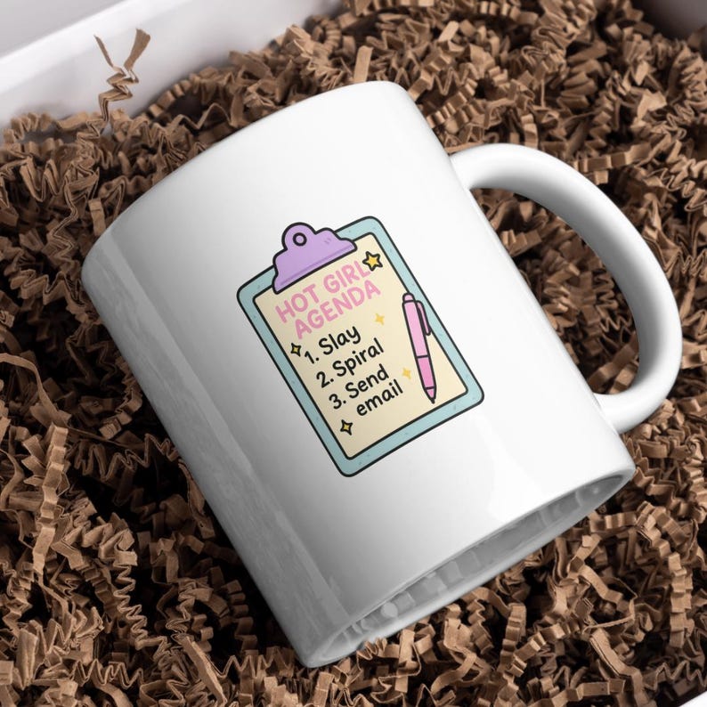 May include: White ceramic mug with a handle, featuring a colorful design. The design includes the text "HOT GIRL AGENDA" above a list: "1. Slay, 2. Spiral, 3. Send email." A pink pen illustration is next to the list.