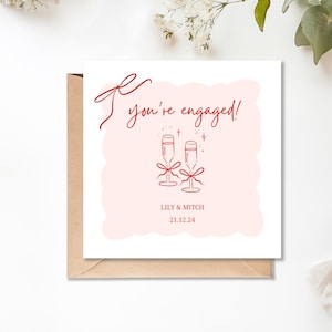 May include: A square engagement card with a pink background and red text that reads "You're engaged!". Two champagne glasses with bows are below the text. The names "Lily & Mitch" and the date "21.12.24" are also on the card.