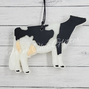 May include: White and black cow silhouette ornament with a light brown spot. The ornament is made of resin and has a string for hanging.