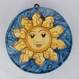 Może przedstawiać: Ręcznie malowany ceramiczny wiszący na ścianie słońce. Słońce ma uśmiechniętą twarz z żółtymi promieniami i niebieskim tłem z dekoracyjnymi wirami. Czarna metalowa pętla jest przymocowana do zawieszenia.