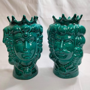 May include: Two ceramic green vases shaped like human heads with ornate details. The vases are both wearing crowns and have facial features.