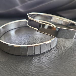 May include: Two silver-colored bangles. One bangle has a series of rectangular indentations, while the other has a smooth, geometric design with a black accent. The bangles are displayed on a dark surface.