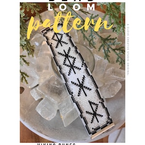 Puede incluir: Un brazalete de cuentas blanco y negro con un patrón de runas vikingas. El brazalete está sobre una superficie blanca con cristales transparentes.