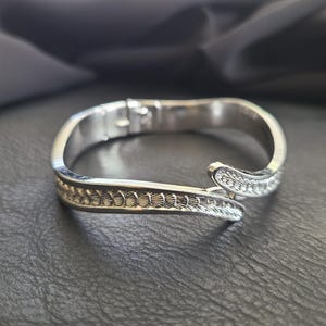 May include: A silver-colored hinged bracelet with a decorative pattern. One side features a series of small, circular designs, while the other is adorned with sparkling, clear stones. The bracelet is open, showcasing its clasp mechanism.