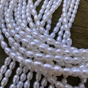 May include: A collection of lustrous, white oval-shaped pearls. The pearls are strung together, creating multiple strands. The pearls have a smooth, reflective surface, and the overall effect is one of elegance and sophistication. The background is a dark, wooden surface.