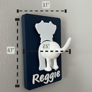 May include: A white dog-shaped hook with a blue background and the name "Reggie" printed on it. The hook measures 4.7 inches tall and 3.1 inches wide.